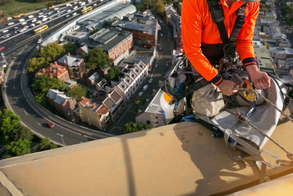 Worker caring for commercial roof using abseiling equipment.
